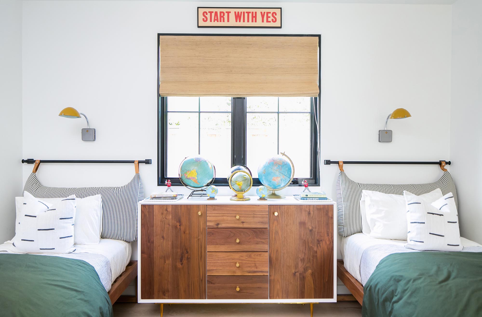 A children’s room with smartly wooded cabinetry surrounded by matching twin beds sports unique headboards composed of oversized lumbar ticking patterned pillows attached to the wall with leather straps.