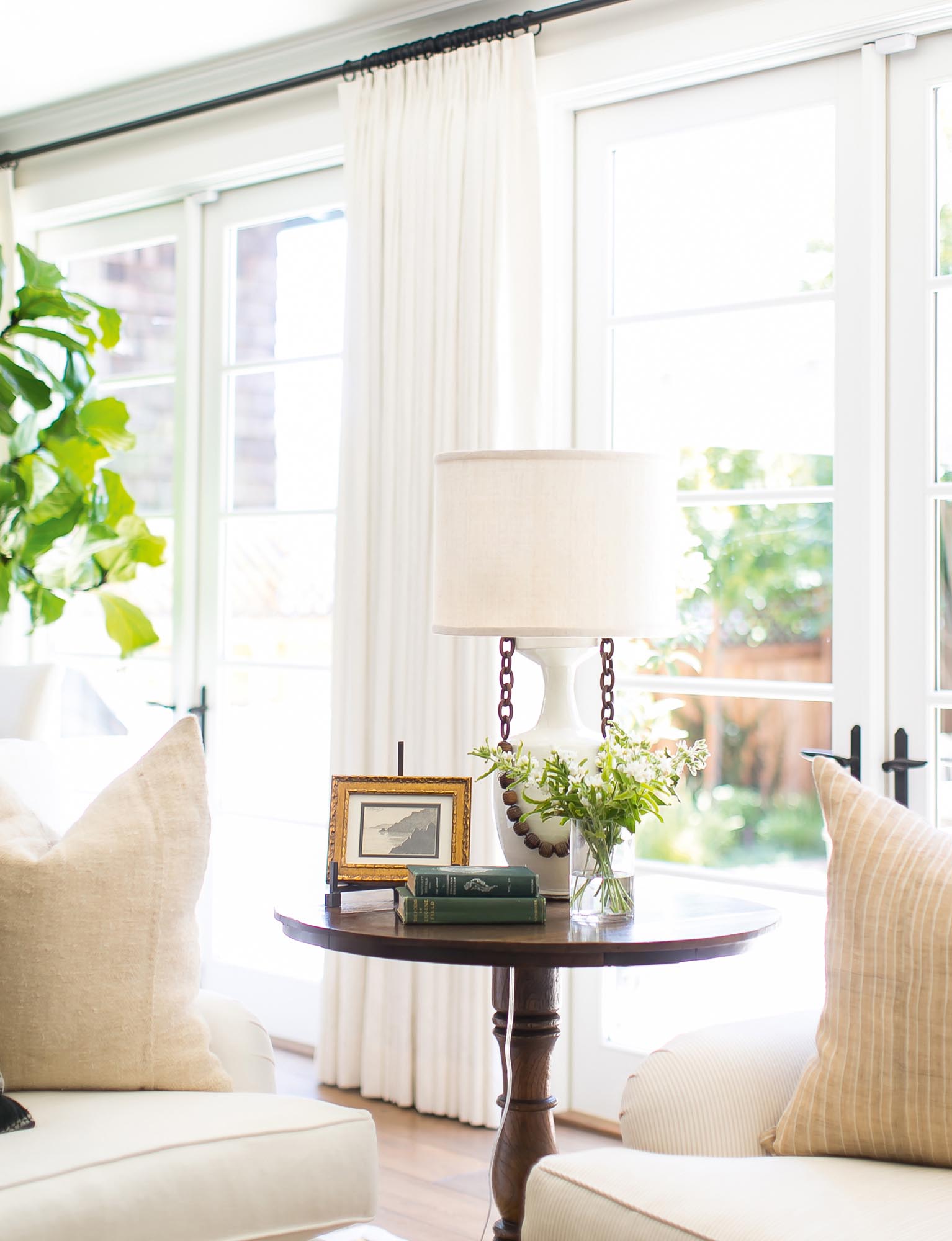 Photo of a tall side table holds vintage books, a small framed artwork, a white lamp with chain and wood bead details, and a simple vase of flowers.