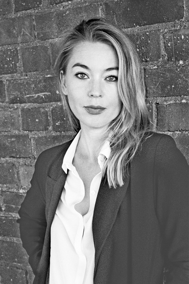 Author photo of Karolina Ramqvist. She is wearing a black blazer and a white button down shirt underneath. She has medium length, blond hair that is draped over her left shoulder. She is looking directly at the camera. In the background is a brick wall.