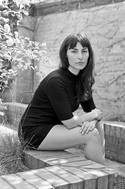 Author photo of Saskia Vogel. She is wearing a black mockneck shirt with sleeves that go to her elbows and black short shorts. She has long dark hair that hangs loosely over her left shoulder. She is sitting on a brick ledge in the middle of a courtyard, turned slightly to the right. She is looking straight at the camera with a serious expression.