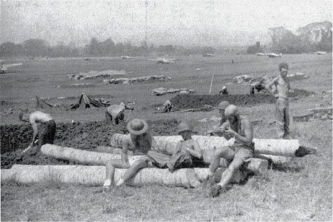 In late September 1942, VF-5 pilots (l to r) Cash Register, Junior Kleinmann, and George Morgan rest near the VF-5 ready tent, while Marines emplace 75-mm pack howitzers. VF-5 F4Fs are parked at the east end of Henderson Field. (Capt. H. W. Crews)