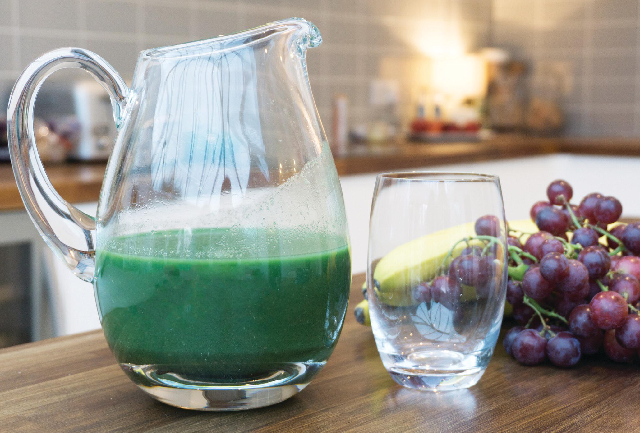 Photo displaying a pitcher of drink, an empty glass, a bunch of grapes, and bananas on a table.