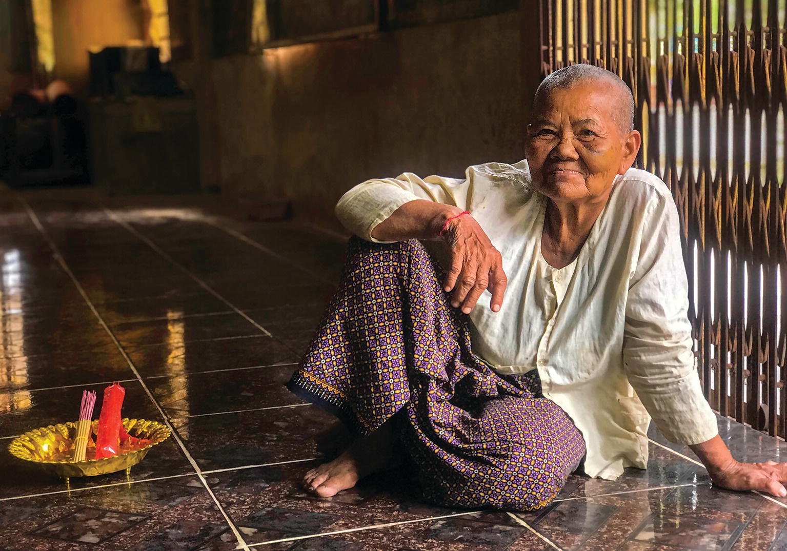 Photo displaying an old woman sitting on the floor. A bunch of incense in a bowl is observed at the left side.