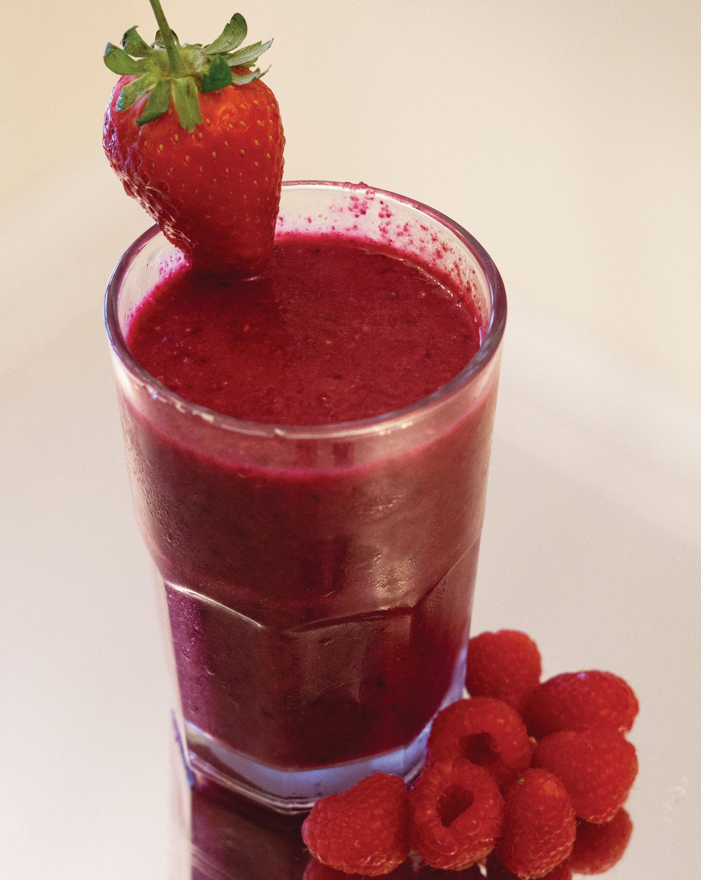 Photo displaying a glass smoothie. A strawberry is placed at the lid of the glass. A heap of raspberries is placed beside the glass.