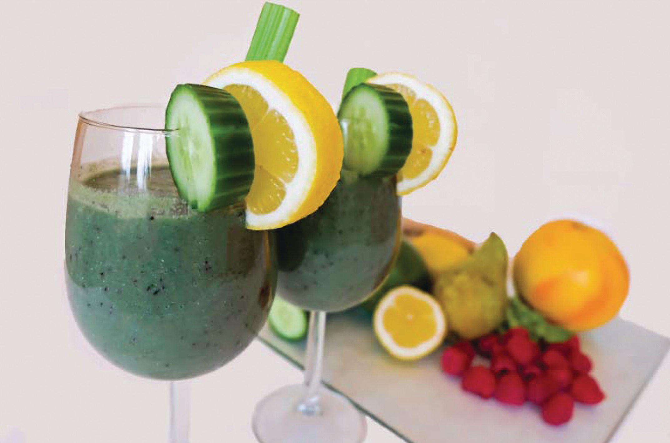 Photo displaying two glasses of cucumber juice in the foreground and a sliced orange, few raspberries, cucumber, and other fruits and vegetables at the background.