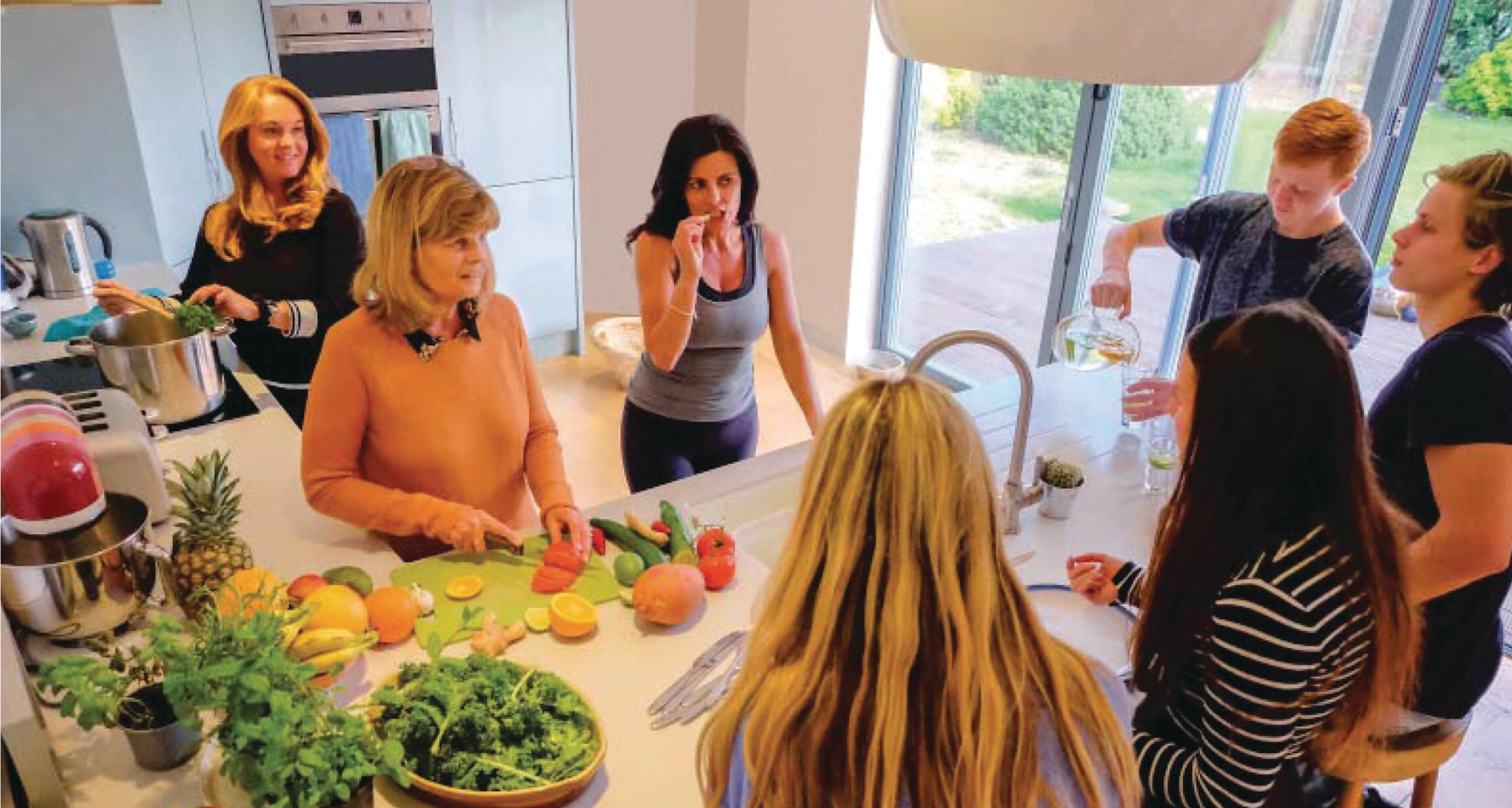 Photo displaying a group of seven individuals in a kitchen, with one of them slicing fruits and vegetables.