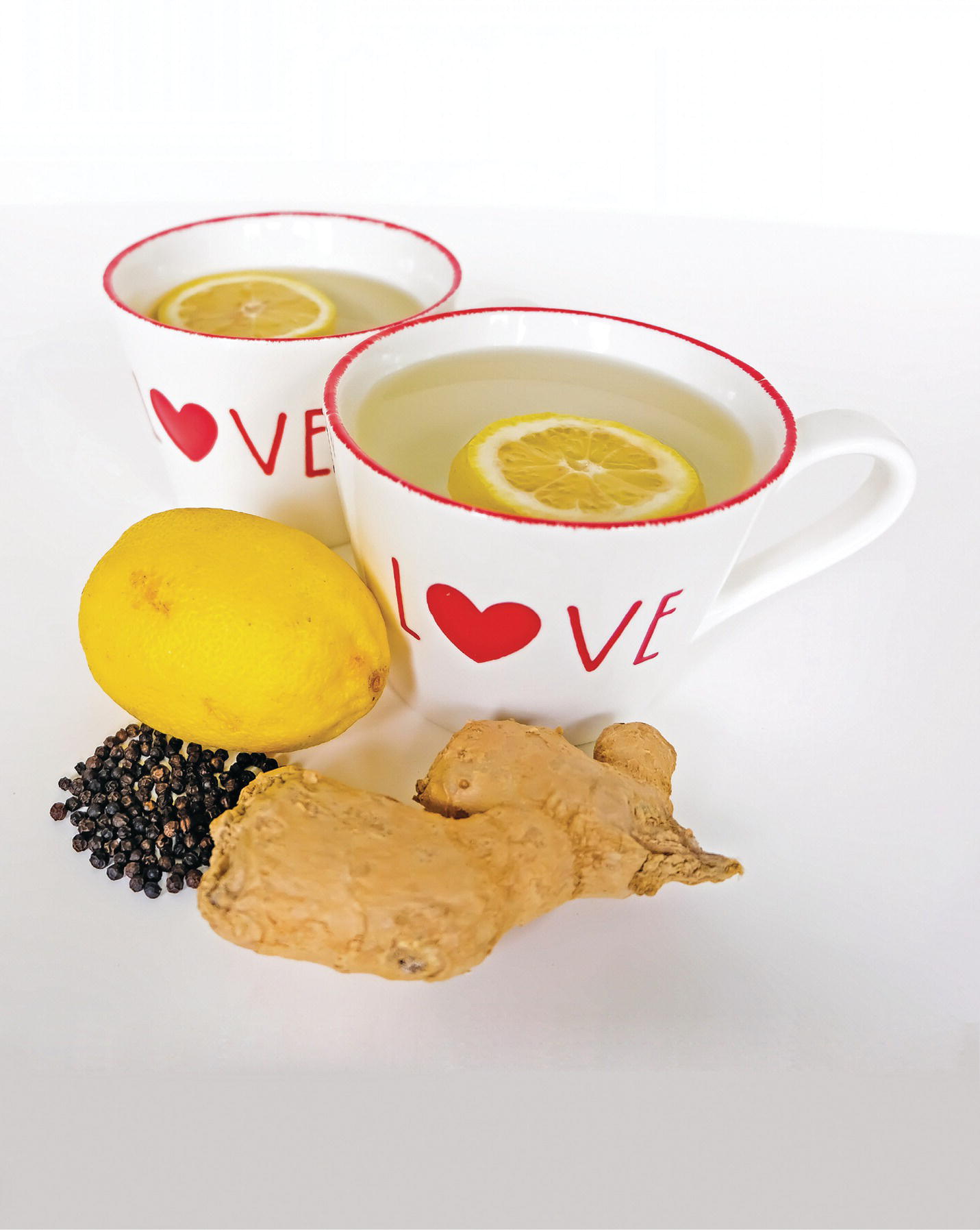 Photo displaying a lemon, ginger, black pepper seeds, and two cups of water with sliced lemon.