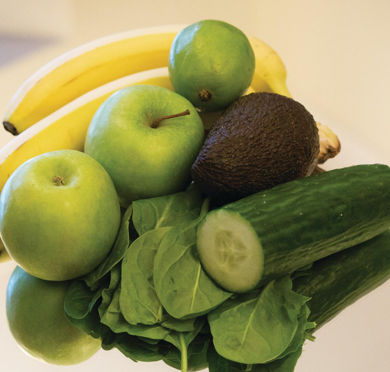 Photo displaying two bananas, two green apples, a lime, an avocado, spinach leaves, and a cucumber.