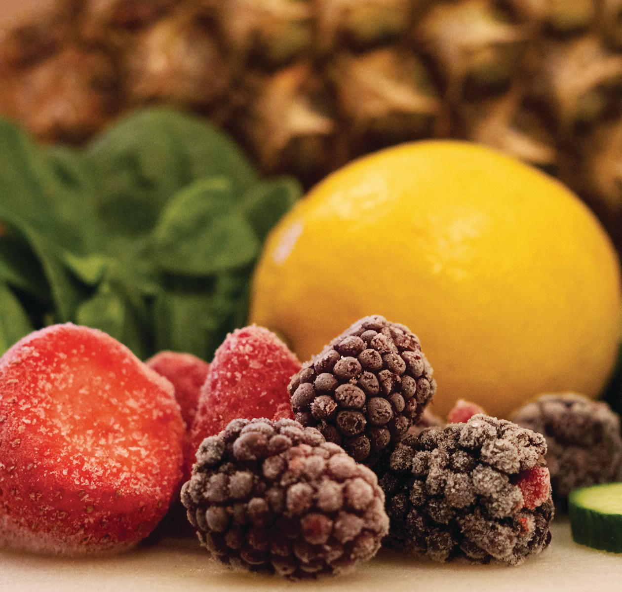 Photo displaying a pineapple, a handful of green leaves, a slice of cucumber, a whole lemon, frozen raspberries, and frozen strawberries.