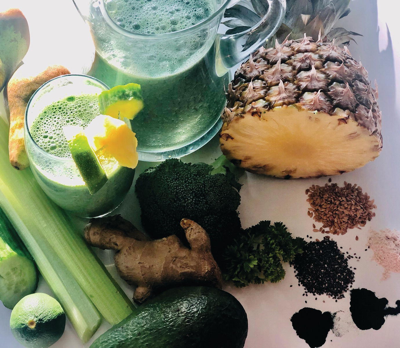 Photo displaying a pitcher and a glass of tropical goodness smoothie. A sliced pineapple, ginger, broccoli florets, avocado, celery sticks, sliced cucumber, parsley, flax seeds, and chia seeds are also observed.