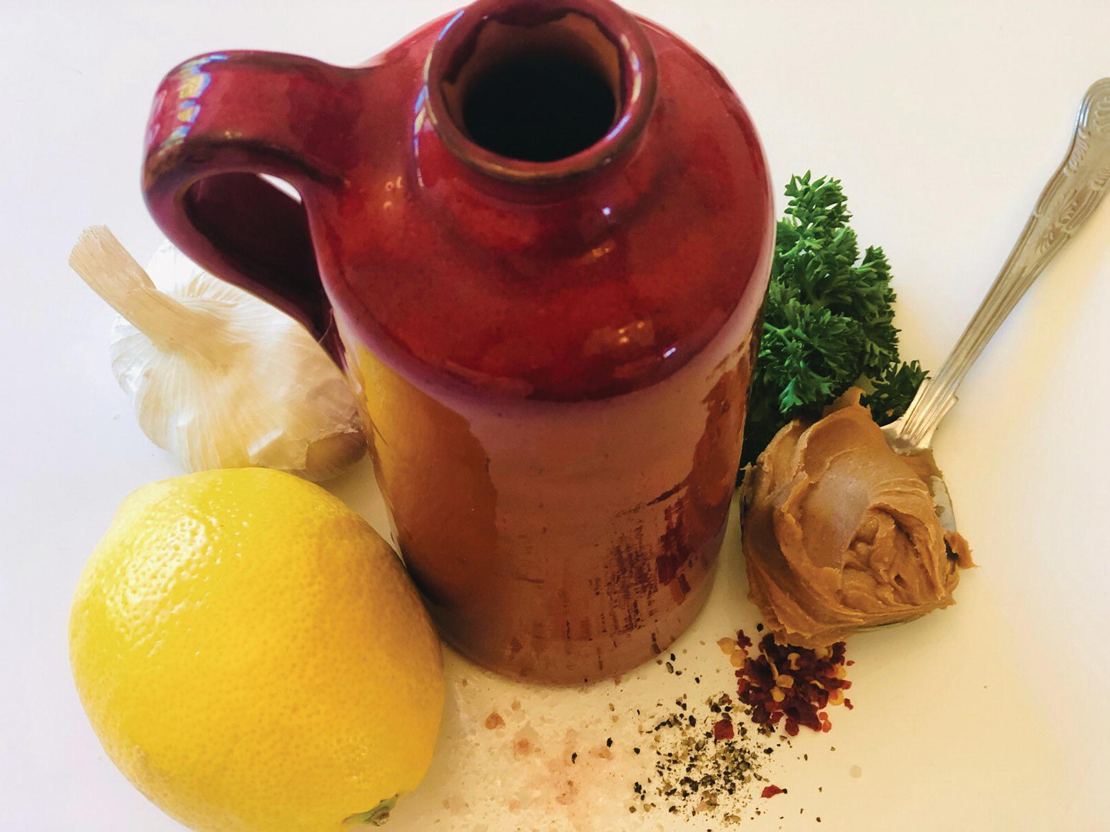 Photo displaying a jar, a whole lemon, a bulb of garlic, a teaspoon of almond butter, fresh parsley, Himalayan salt, black pepper, and chili flakes.