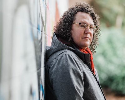 A man with long curly hair wearing a grey jacket and red scarf. He is standing in front of a brick wall with graffiti on it.