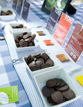 Small square bowls filled with chocolate wafers, each bowl sitting in front a small colorful sign indicating what kind of chocolate it is. The bowls sit in a long row on a table covered in a blue-and-white checkered tablecloth.