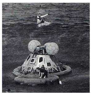 As Odyssey is kept afloat in the South Pacific Ocean after it splashed down at 12:07 P.M. (CST) on April 17, 1970, astronaut John L. Swigert is lifted in a Billy Pugh net to a helicopter, where Fred W. Haise has already been taken. James Lovell waits in a life raft with a U.S. Navy underwater demolition swimmer, while other navy team swimmers involved in the recovery operation are in the water.