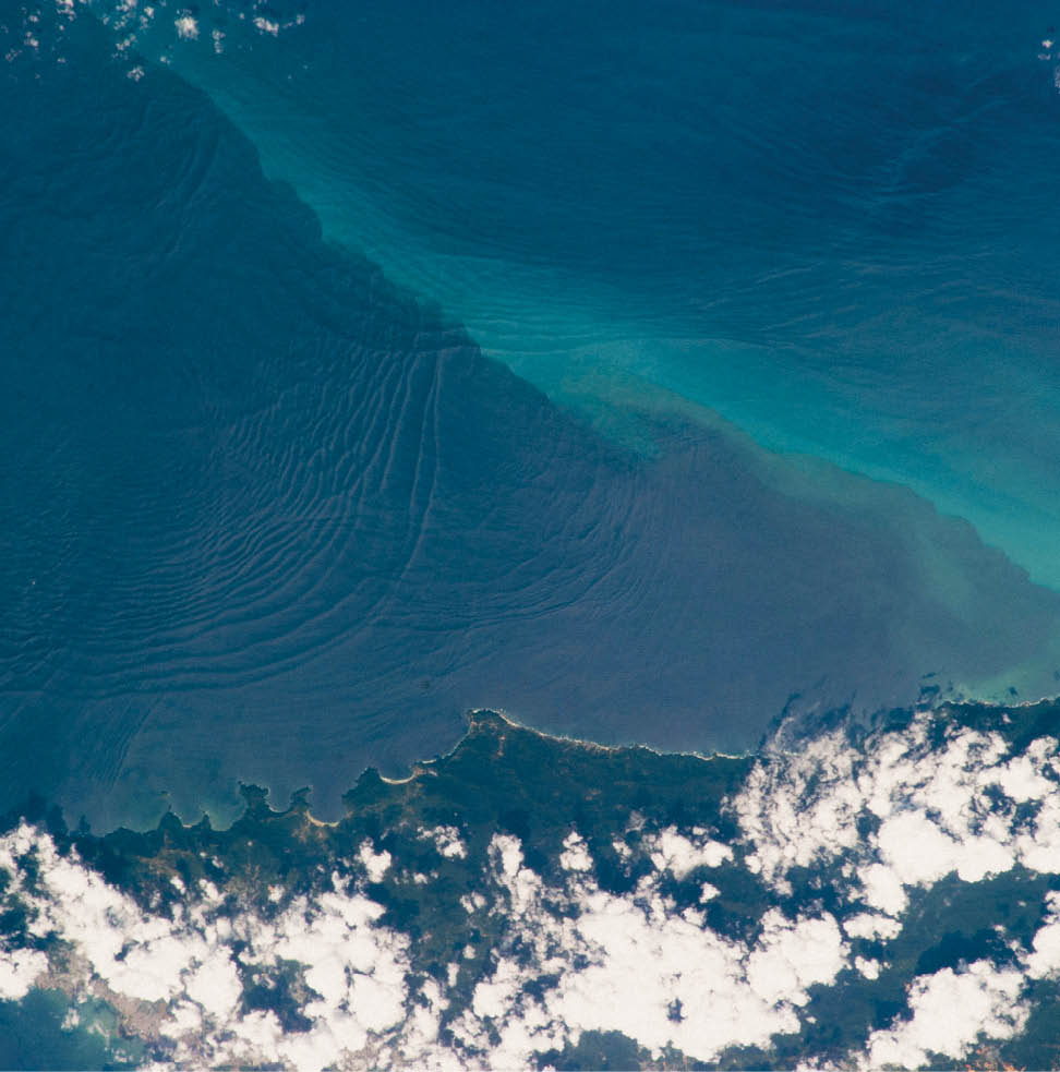 Internal waves are the surface manifestation of slow waves that move tens of meters beneath the sea surface. These waves beneath the waves produce enough of an effect on the sea surface to be visible from space when they are enhanced by the reflection of sunlight, or sunglint, back toward a camera. This January 2013 photograph from the International Space Station shows at least three sets of internal waves interacting. The most prominent set (top left) shows several waves moving from the northwest due to the tidal flow toward the north coast of Trinidad. Two less prominent sets can be seen further out to sea. All of these internal waves are probably caused by the shelf break near Tobago. The shelf break is the step between shallow seas (around continents and islands) and the deep ocean. It is the line at which tides usually start to generate internal waves.