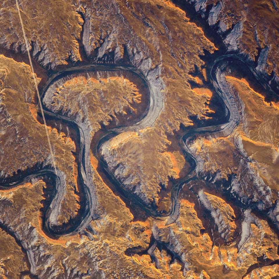 This section of the Green River canyon in eastern Utah is known as Bowknot Bend because of the way the river doubles back on itself.  In this January 2014 photograph taken from the International Space Station, the Green River appears dark because it lies in deep shadow, 300 meters (1,000 feet) below the surrounding landscape. The yellow-tinged cliffs that face the rising Sun give a sense of the steep canyon walls. The straight white line across the scene is the contrail from a jetliner.