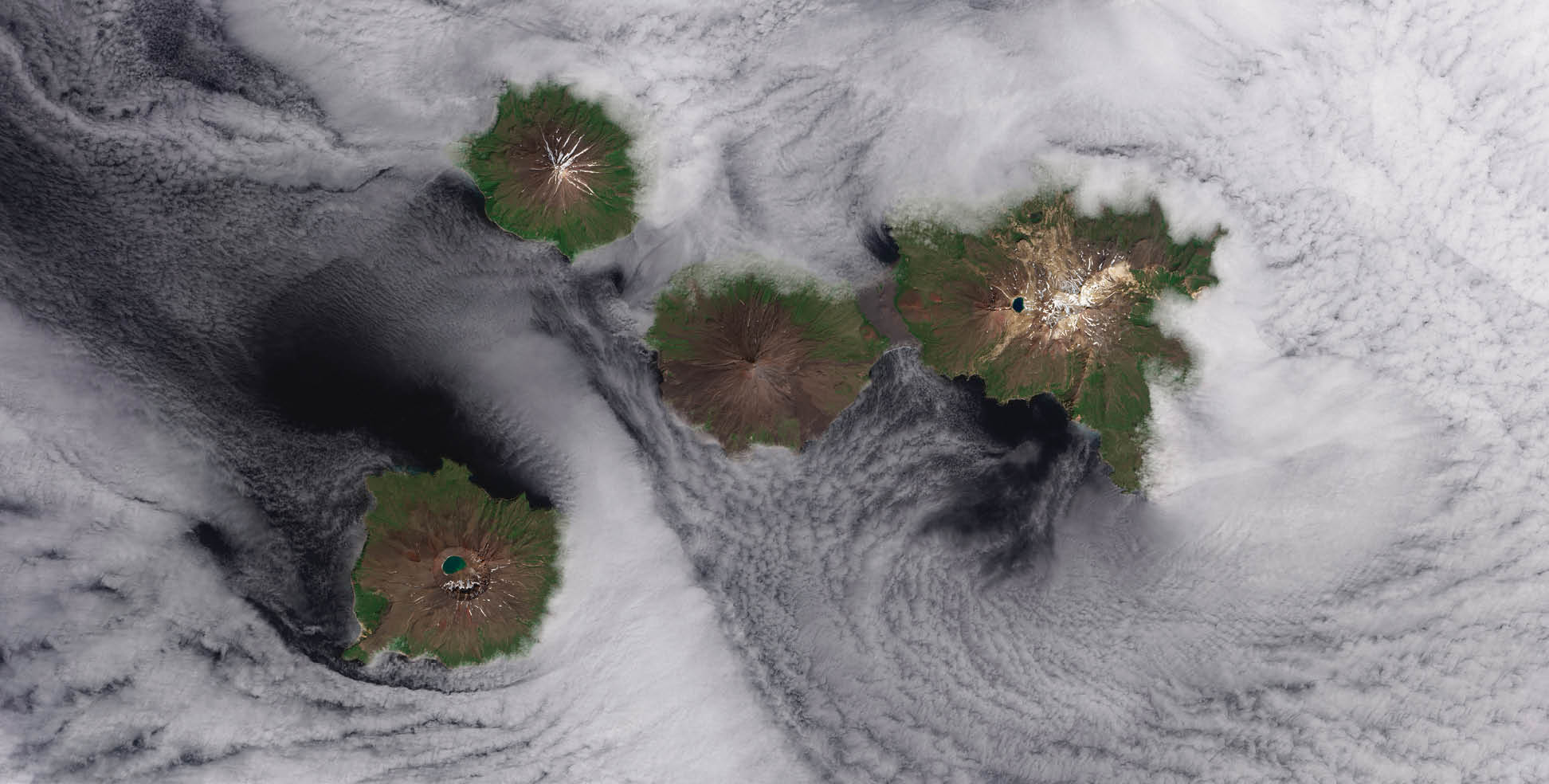 They are called the Islands of the Four Mountains. Part of the Aleutian Island chain, these peaks are actually the upper slopes of volcanoes rising from the seafloor: Carlisle, Cleveland, Herbert, and Tana. Standing in one of the most remote reaches of the world, these volcanoes have scarcely been studied. Satellite sensing makes that easier, as this Landsat 8 image from June 2013 shows. Herbert Island (right) is dominated by a symmetrical stratovolcano that stands in its center. The remote island has scarcely been studied, and there are no records of eruptions there. The 2-kilometer-wide summit caldera include a lake of meltwater, remnants of the snow that covers the peak for most of the year. The straight-down (nadir) satellite view can make it difficult to determine which part of the landscape stands taller than the other, a phenomenon known as relief inversion.