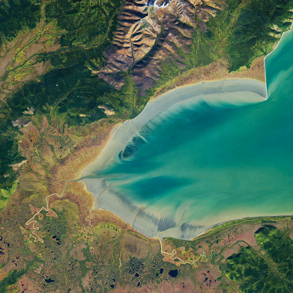 Onshore, freshwater streams around Russia’s Ulbanskiy Baymeander into marshlands and gently sloped mud flats. The marshes around the bay are dotted with small bodies of water. These are likely thermokarst lakes—pools of water that fill in depressions in the land surface as permafrost melts. Farther inland, the incline becomes steeper and the landscape darker with the greens of pine-covered slopes. A lighter green band (left side) indicates deciduous (leaf-shedding) trees that have begun to turn color. 