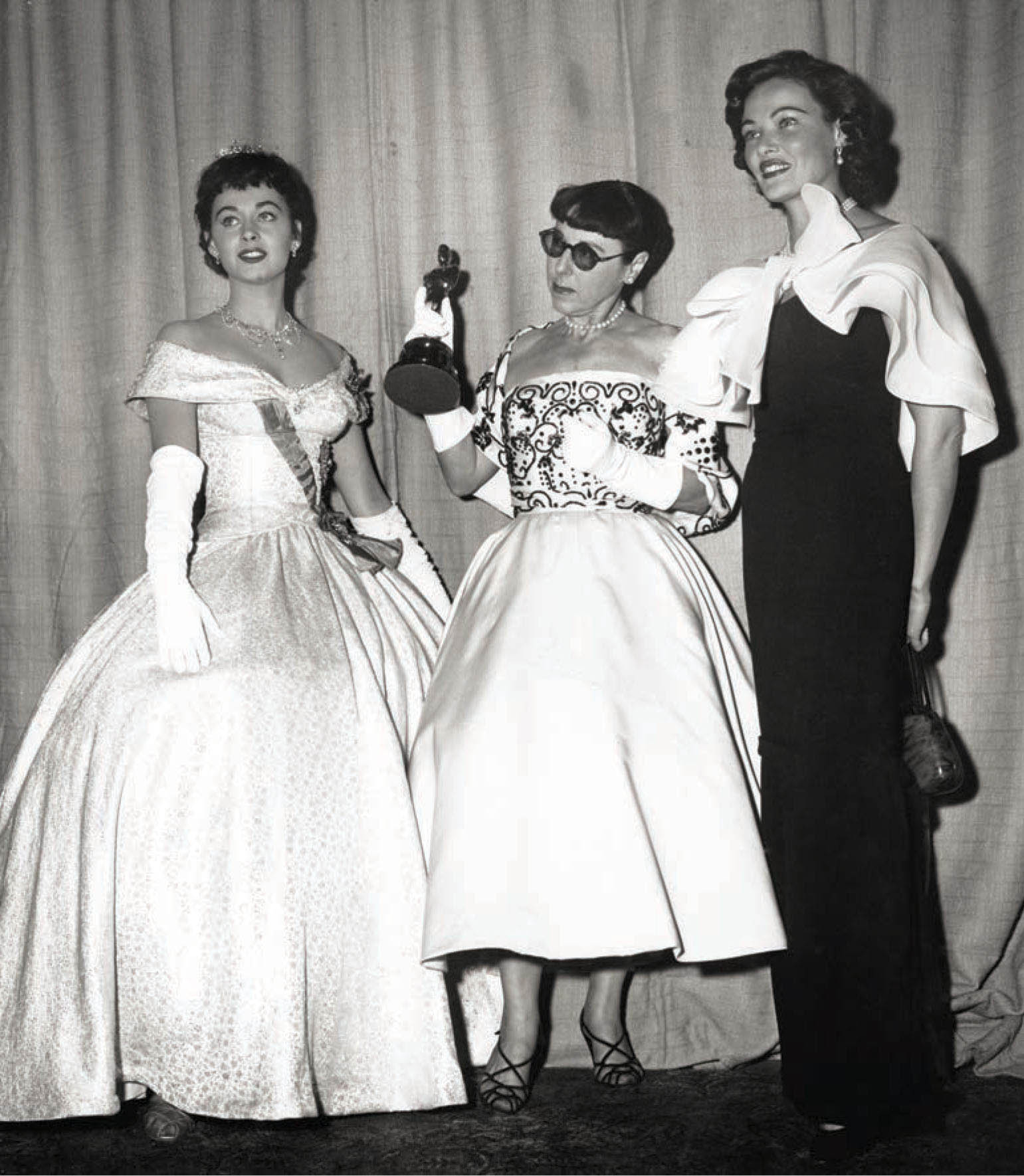 (l-r) Marla English wearing Audrey Hepburn’s gown from Roman Holiday (1953), stands next to Edith Head and actress Gene Tierney. Edith is holding the Oscar she won for designing the film.