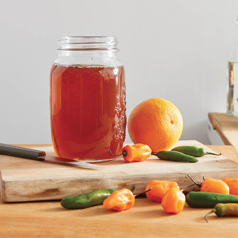 A jar of honey with an orange and peppers on a cutting board.