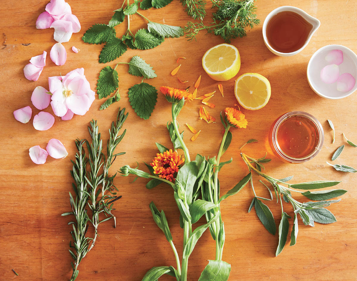 A spread of fresh picked herbs, flowers, lemon, and jars of honey.