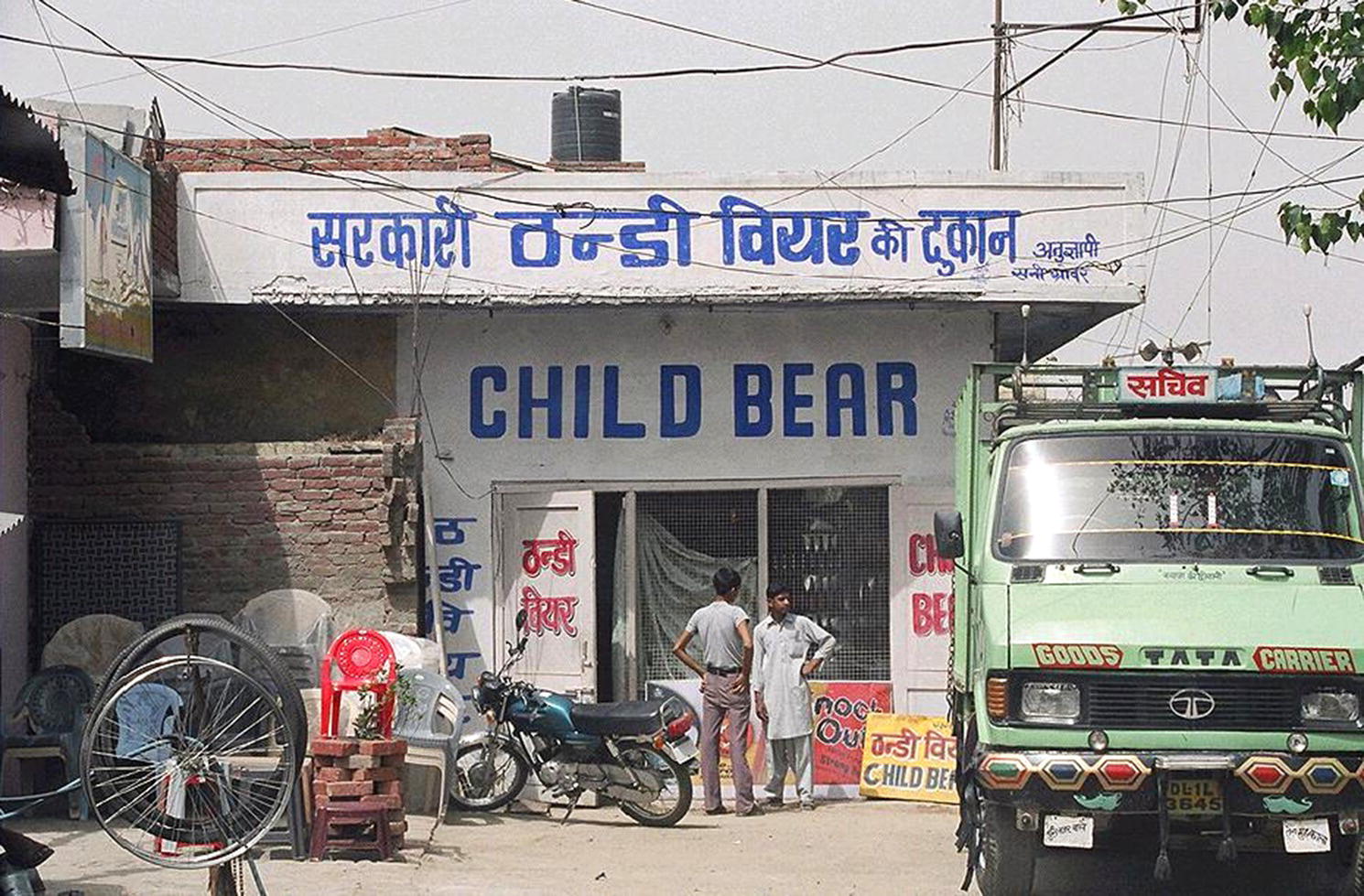 Photo displaying an Indian shop with a sign “Child Bear.” Two men and a motorcycle are observed in front of the shop. A truck is observed at the right side.