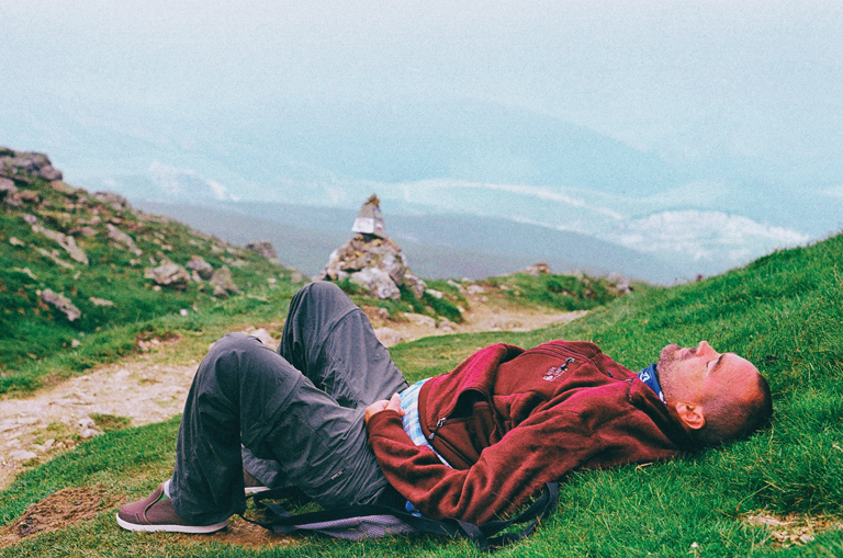 Justin lies on a grassy hillside, his eyes closed. In the distance are mountain peaks.