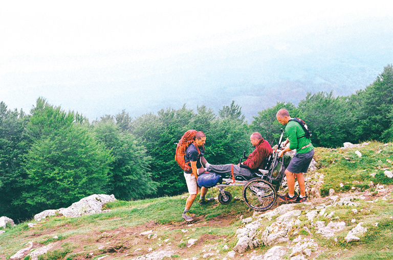 Patrick and Joe lift Justin in his wheelchair over a rocky spot in the unpaved mountain path.