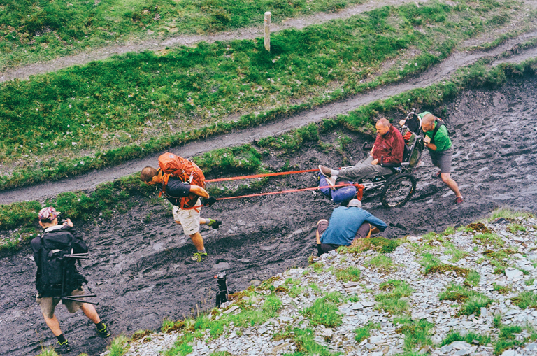 Patrick pulls Justin in his wheelchair, while Joe pushes from behind. The trail is a wide muddy trench with puddles of standing water.