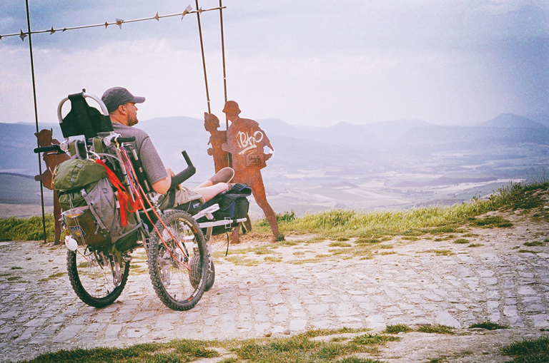 Justin looks out over a valley from the top of a hill. Beside him are three life-size metal silhouettes of pilgrims walking.