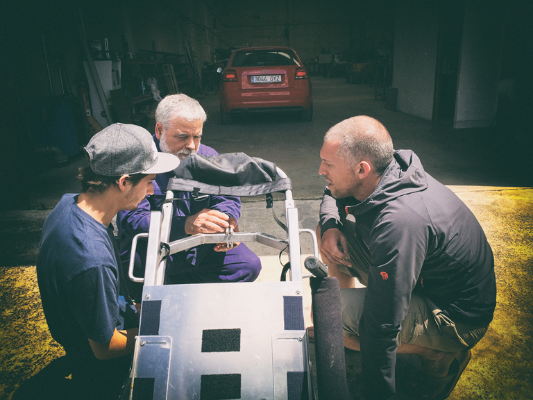 Justin, a local welder, and Patrick sit on the ground around the wheelchair. The welder holds a metal bracket in place against a bar of the wheelchair.