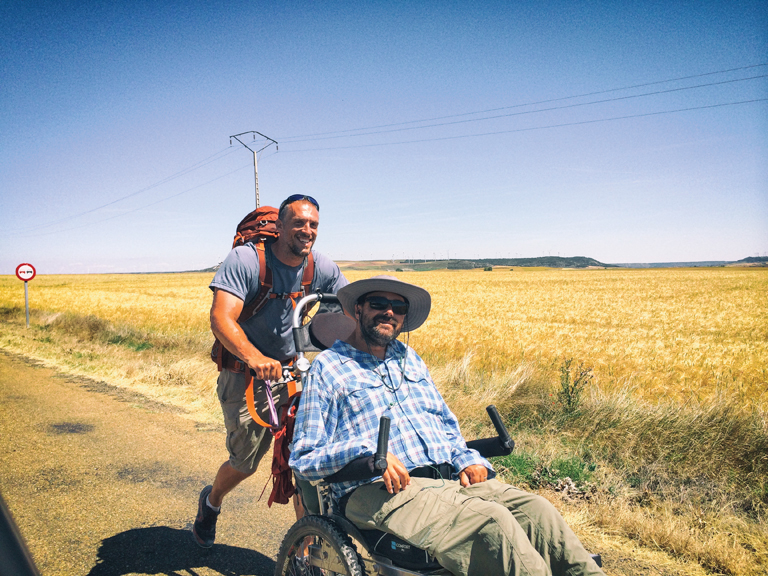 Patrick pushes Justin in his wheelchair on a level asphalt path beside a golden field. The sun is bright and the sky is cloudless.