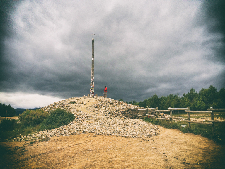 Patrick faces a tall pole with a cross at the top. The pole stands on top of a mound of rock and dirt beneath dark clouds.