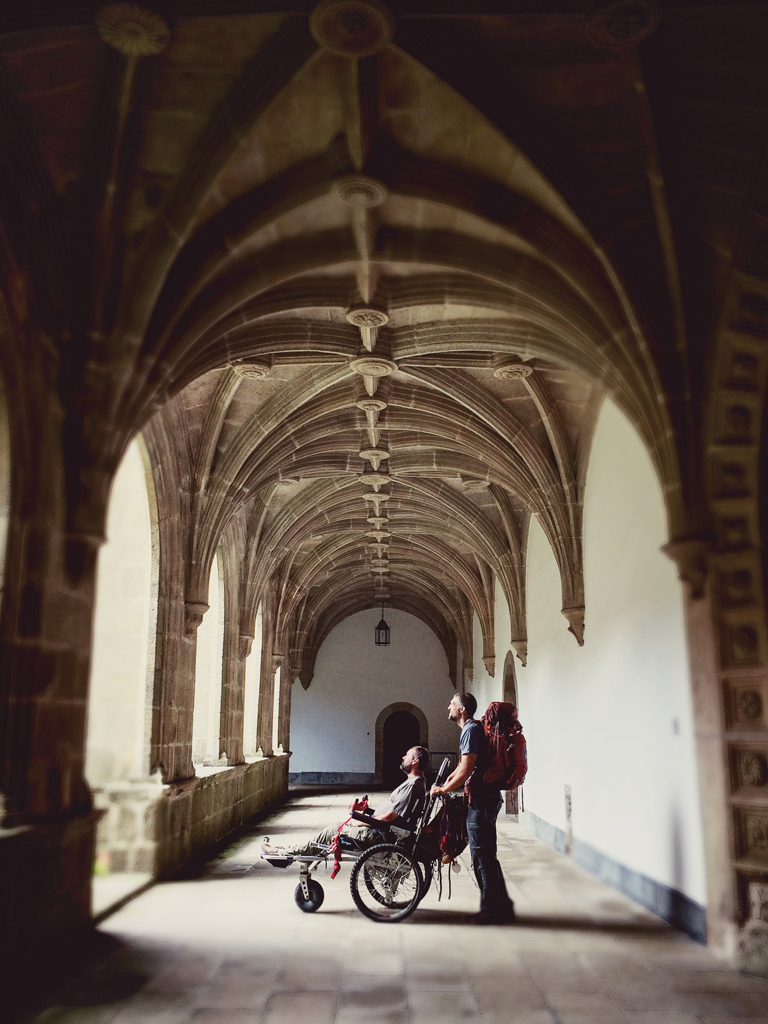 Patrick stands behind Justin beneath vaulted arches. Both are looking out of the arches into the sunlight.