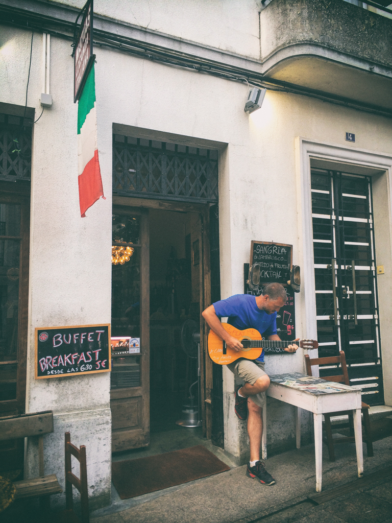 Patrick leans against the exterior wall of a building, playing guitar.