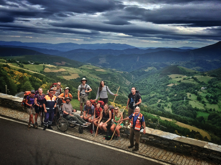 Justin and Patrick pose with thirteen other pilgrims in front of a green valley covered in farm fields and woods.