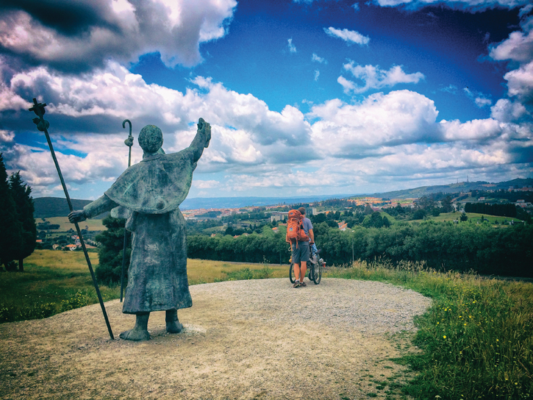 Patrick pushes Justin downhill ahead of us on a gravel trail. In the foreground is the back of a statue that points down the trail with his right hand and holds a walking stick with a cross at the top in his left hand.