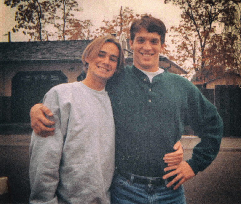 Justin and Patrick pose for a photo in front of a house. They stand side-by-side, each with one arm around the other.