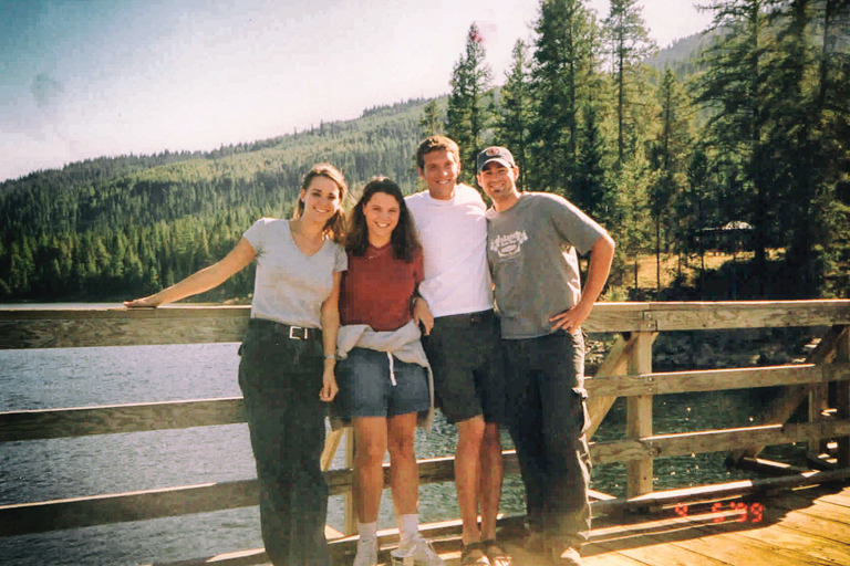 Kirsten, Donna, Patrick, and Justin pose for a photo on a wooden platform overlooking a lake. Pine-covered hills surround the lake.