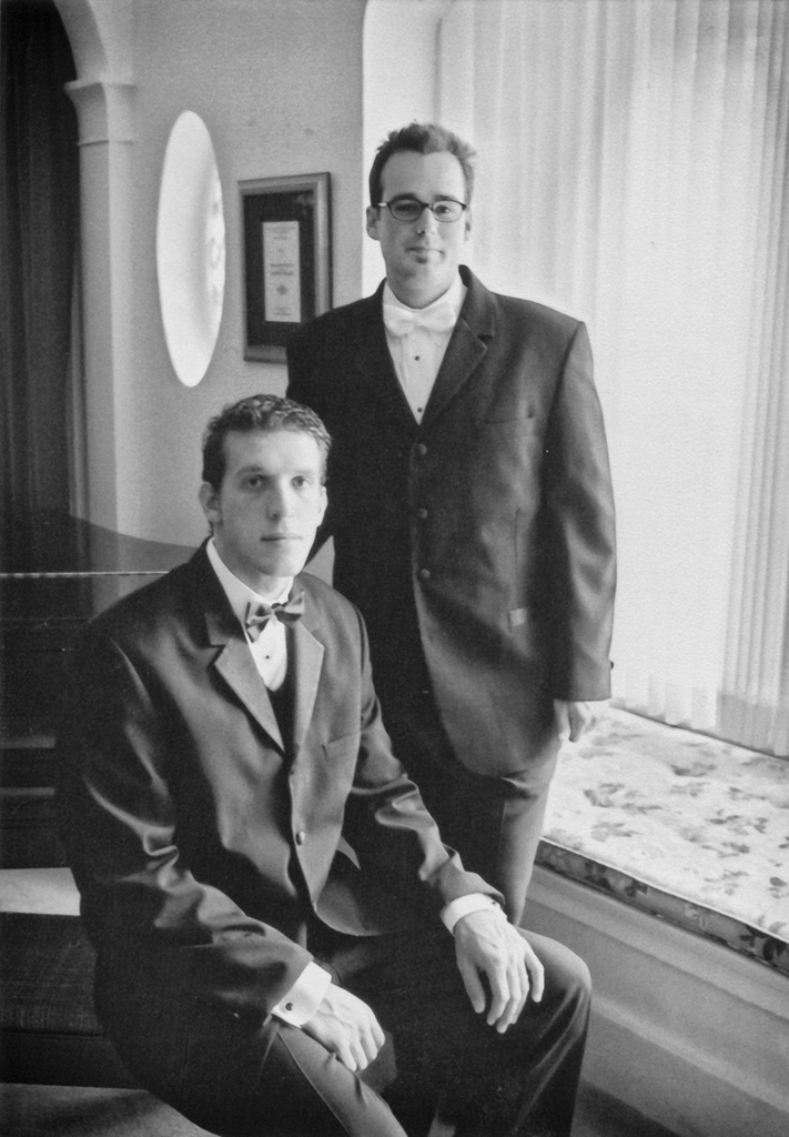 Justin and Patrick pose in tuxedos for a formal photograph. Justin stands behind Patrick, who is seated on a piano bench.