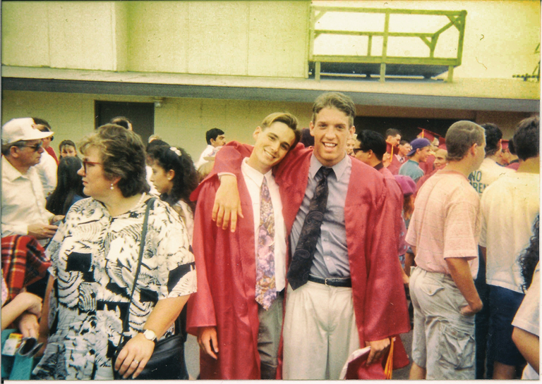 Justin and Patrick pose for a photo in red graduation robes. The robes are open at the front, revealing their button-down shirts, ties, and khakis. They hold their graduation caps in their hands, and Patrick has his arm around Justin’s shoulders.