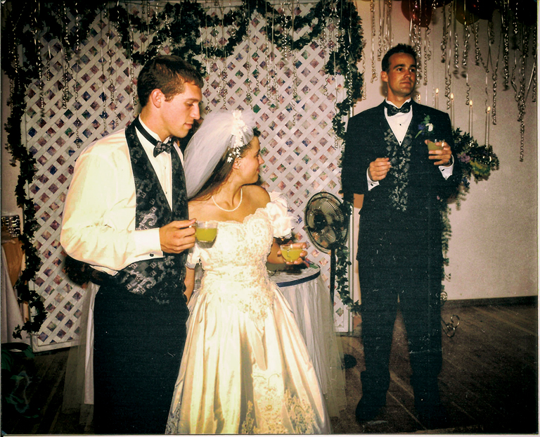 Patrick (in a tuxedo, minus the jacket) and Donna (in a wedding dress) look at Justin, who is holding a microphone. All are holding filled punch glasses.