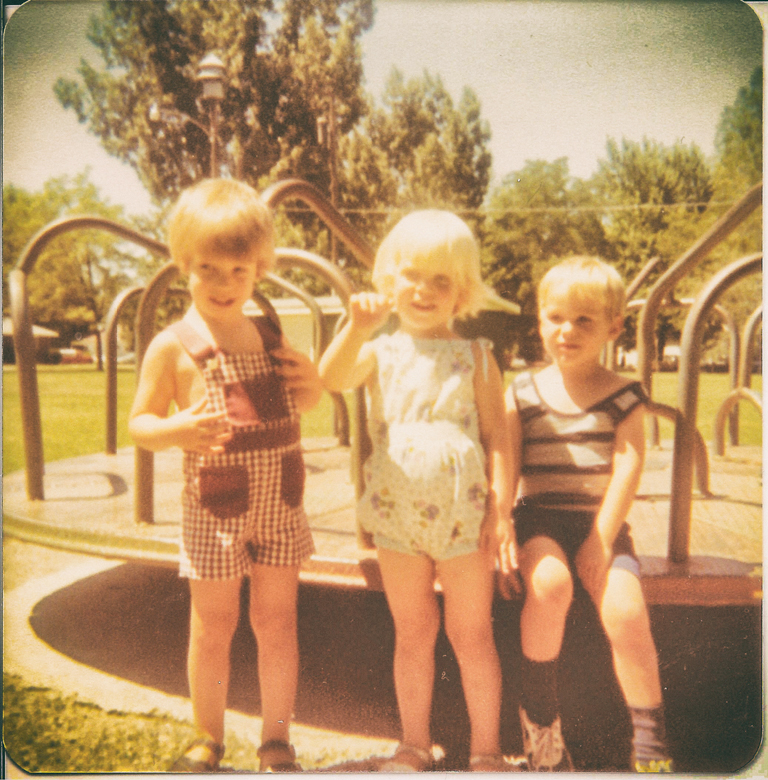 Three children, each about 4 years old, in front of a merry-go-round in bright sunshine.