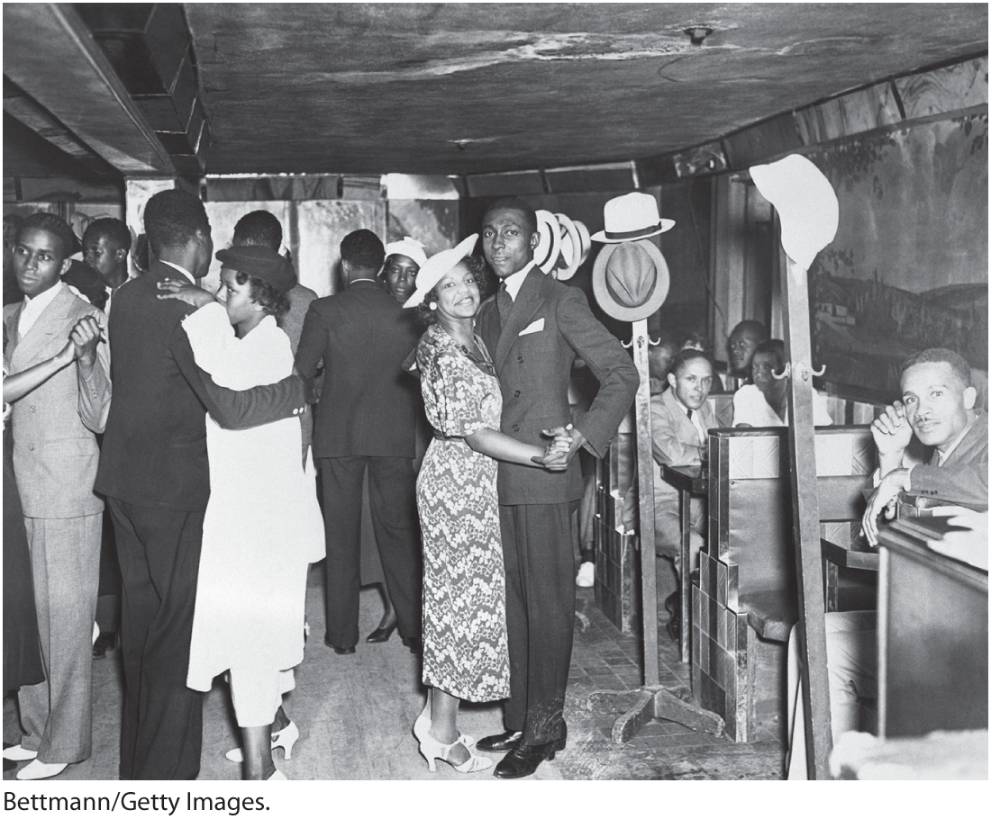 A photo shows men and women dancing at a Harlem club. The men are clad in suits, and the women are clad in dresses and hats.