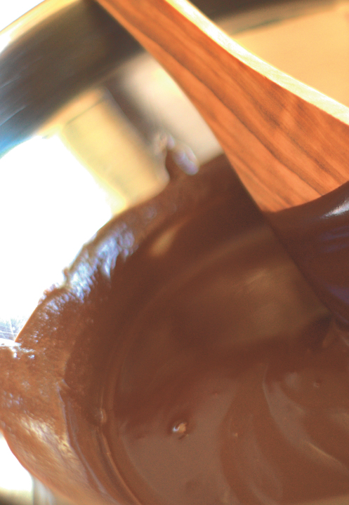 A wooden spoon stirring thin chocolate sauce in a wide metal bowl