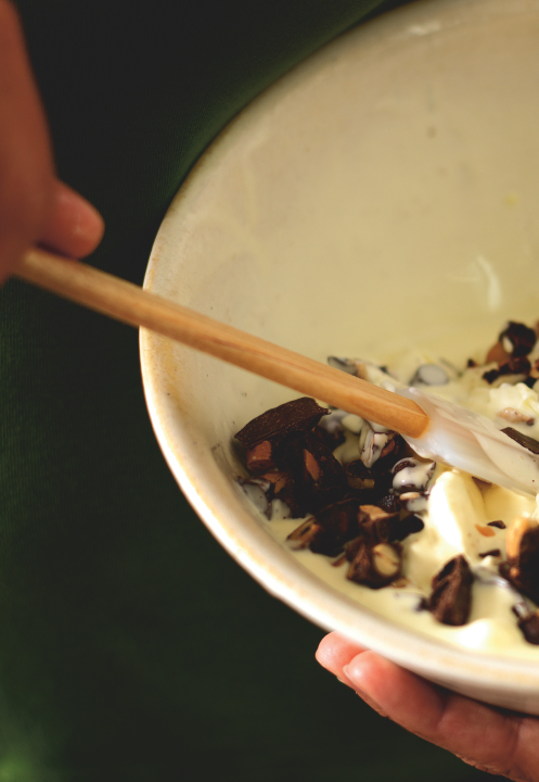 A wooden spoon stirring chopped chocolate and nuts into a large white ceramic bowl filled with smooth vanilla ice cream