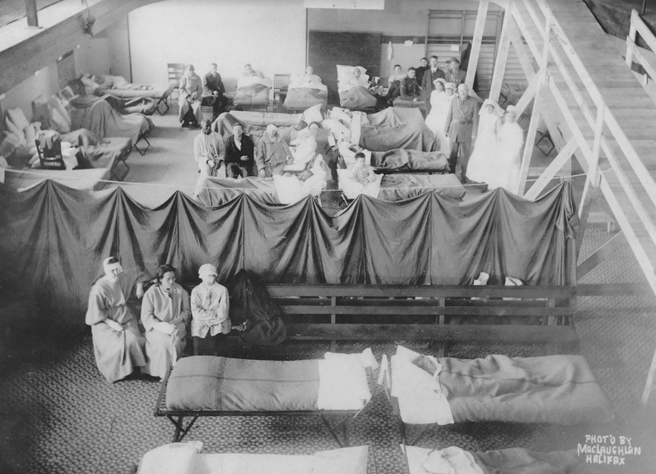 Photograph of a hall in the YMCA, Barrington. Several beds are set in the room, surrounded by staff, patients, and nurses. A few empty beds are also present, which are separated from the rest by curtains.