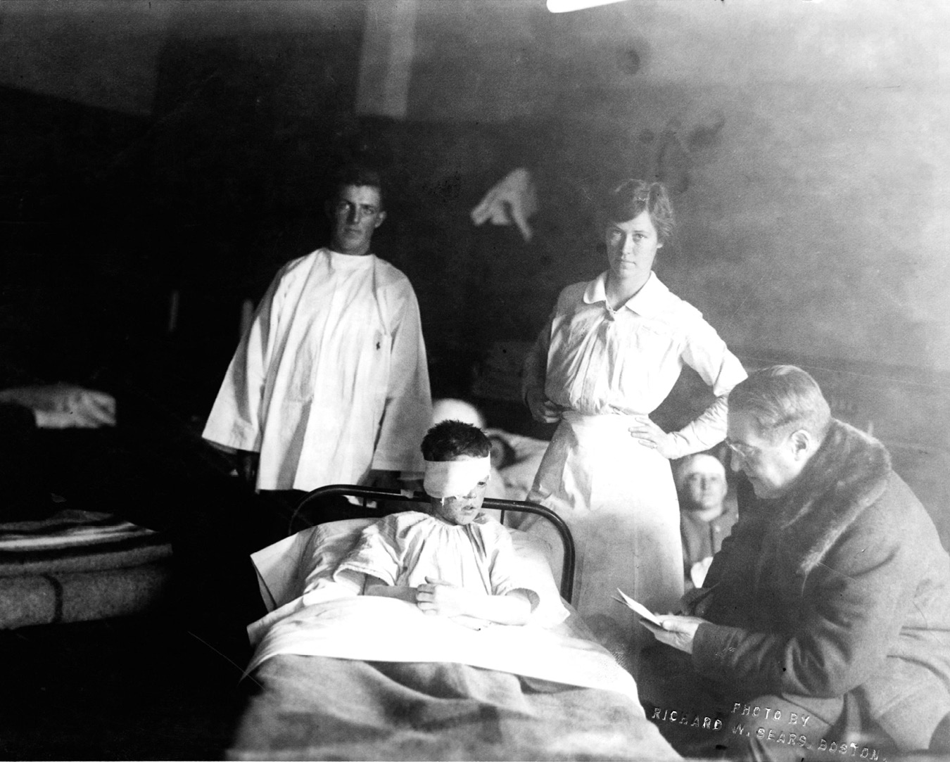 Photograph shows a child with a severe eye injury seated on a bed. The hospital staff standing behind the bed, pose for the photograph. To the right of the bed is Abraham C. Ratshesky, looking at the piece of paper in his hand.