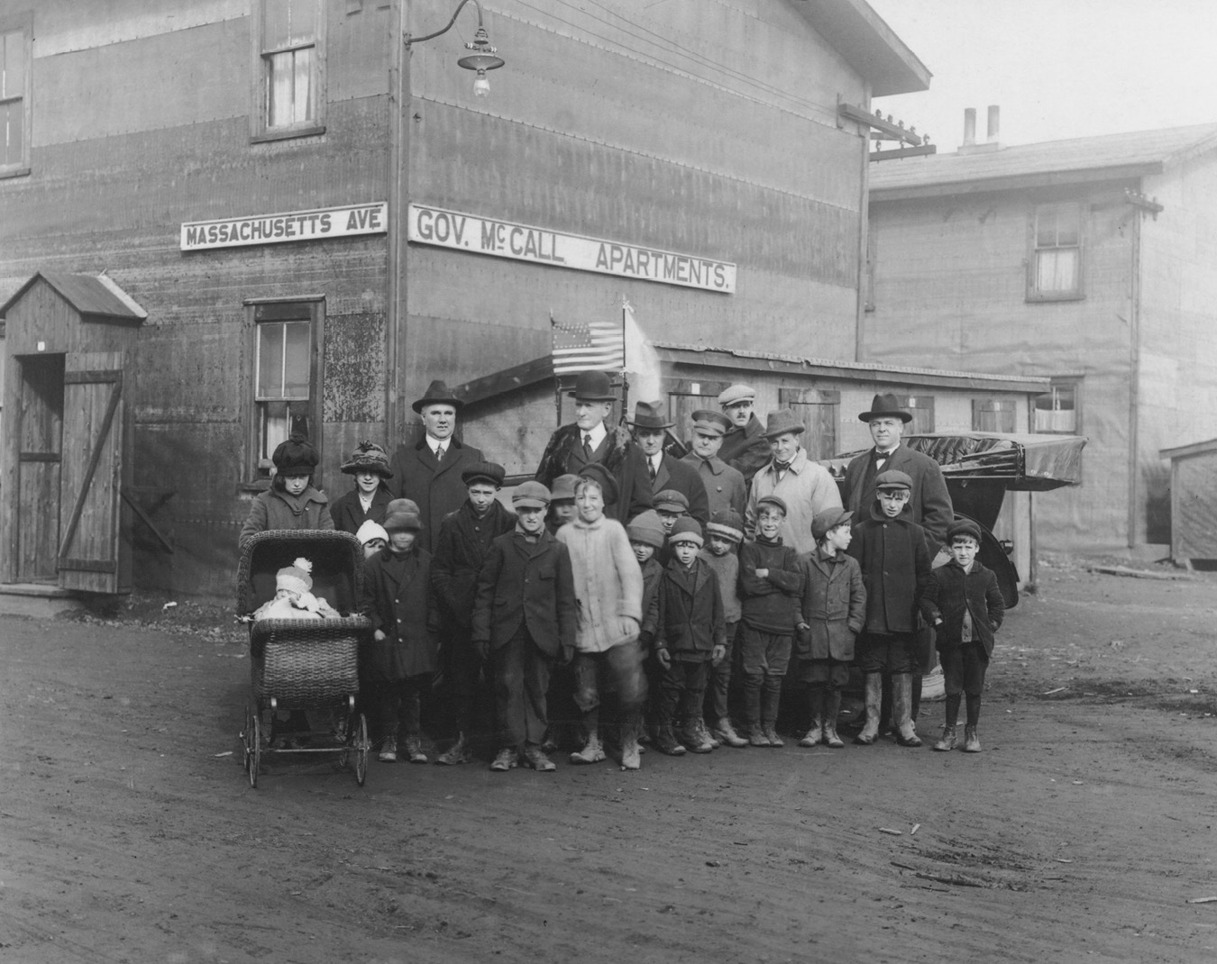 A group of children and a few adults pose for the photograph along with the Governor. On the background is the Governor McCall apartments in Massachusetts Avenue.