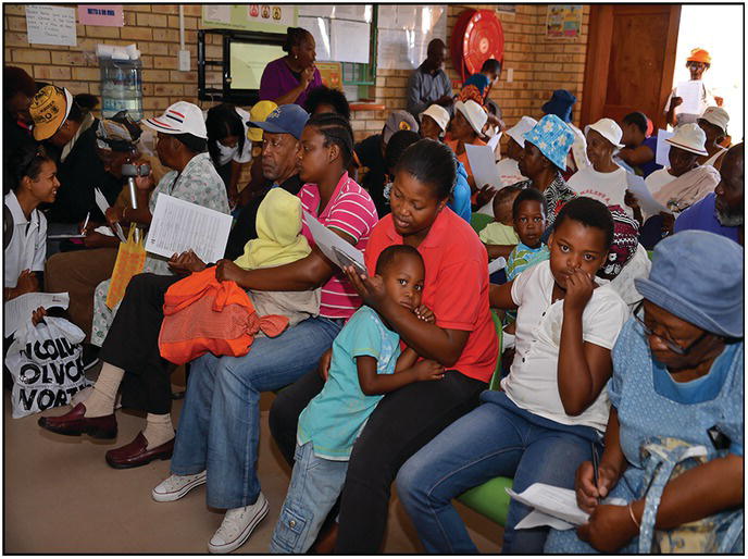 Photo of men and women sitting in queue as they hold, check, and write on their forms.